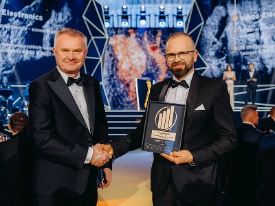 Kuva kahdesta miehestä, jotka ovat pukeutuneet juhlapukuun ja kättelevät toisiaan. Oikealla oleva mies, Maciej Góralski, pitelee kädessään kehystettyä palkintoa, jossa on kultainen tunnus ja joka on merkitty ”Vuoden yrittäjä 2022” -palkinnoksi. Hämärtyneessä taustassa näkyy lava, jolla on abstrakteja geometrisia rakenteita sekä dynaamisia sinisiä ja punaruskeita graafisia kuvioita. Useat vieraat istuvat pöydissä hämärästi valaistussa juhlasalissa.