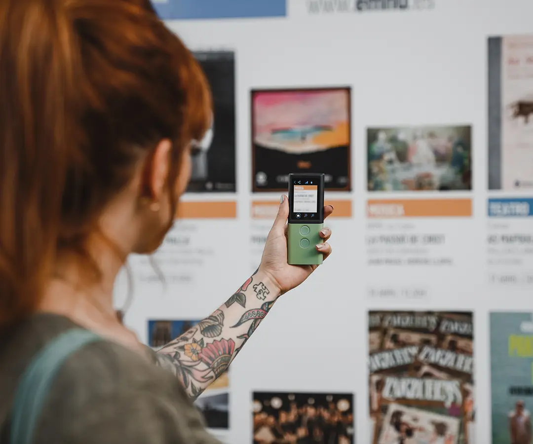 Henkilö, jolla on punaiset hiukset ja kukkatatuointi käsivarressa, pitää kädessään vihreää digitaalista laitetta ja osoittaa sillä kohti näyttötaulua, jossa on erilaisia julisteita ja tapahtumatietoja.