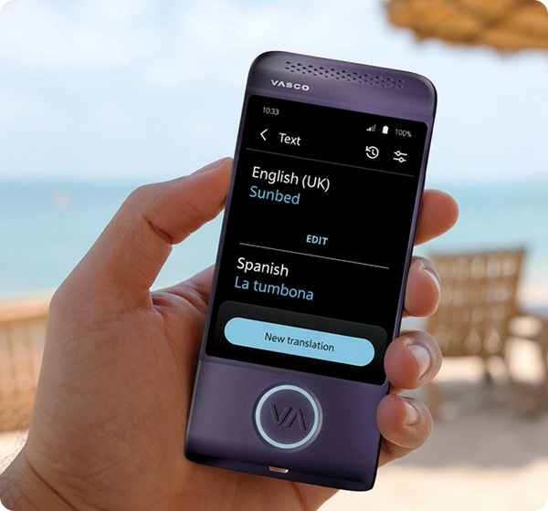 Lähikuva kädestä, joka pitelee violettia Vasco-käännöskonetta. Näytöllä näkyy tekstikäännös englannista (Iso-Britannia) espanjaksi, jossa sana ”Sunbed” on käännetty muotoon ”La tumbona”. Taustalla on valoisa, aurinkoinen rantamaisema, jossa meri, kultainen hiekka, useita aurinkotuoleja ja olkinen rantavarjo näkyvät pehmeästi sumennettuna. 