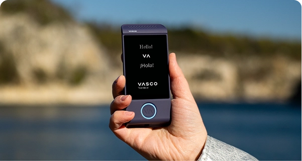 Lähikuva kädestä, joka pitelee violettia Vasco-käännösohjainlaitetta kirkkaalla ulkotaustalla. Näytöllä näkyy tervehdykset ”Hello!”, ”¡Hola!” sekä ”VASCO Translator” -logo. Taustalla on pehmeästi tarkennettu kuva aurinkoisesta rannikkomaisemasta, jossa on sinistä vettä ja vaaleita kallioita kirkkaan taivaan alla. Laitetta pitävällä henkilöllä on vaaleanharmaa tai hopeanvärinen kuvioitu takin hiha. 