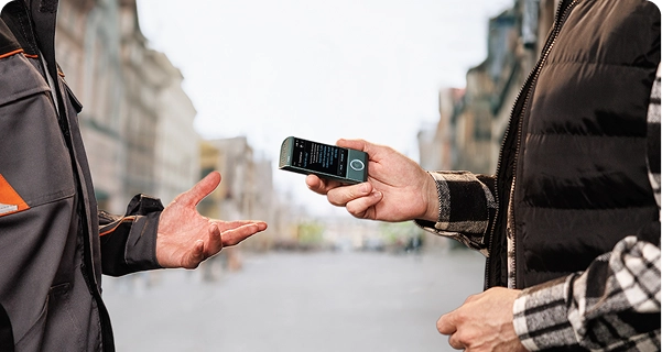 Mies, jolla on yllään musta tikattu liivi ruudullisen paidan päällä, pitää turkoosia Vasco-käännöslaitetta vaakasuorassa toista henkilöä kohti kaupungin kadulla. Toinen henkilö, jolla on yllään harmaa-oranssi työvaate, ojentaa kätensä ikään kuin keskustelun keskellä. Laitteen näytöllä näkyy käännösliittymä, jossa on tekstiä. Taustalla on laaja, hämärtynyt näkymä eurooppalaistyyliselle kadulle, jossa on korkeita rakennuksia kirkkaan, pilvisen taivaan alla. 